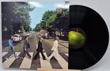 A vinyl record partially pulled from its sleeve, showing the iconic Abbey Road album cover with four men walking across a zebra crossing and a black record disc with a green center label on a light gray background.