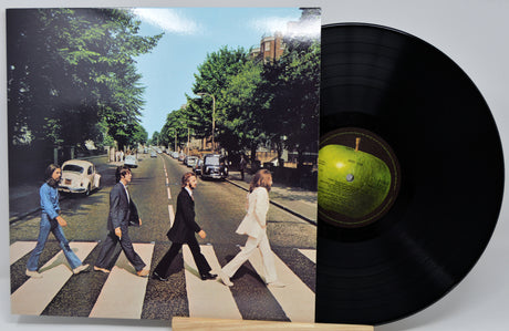 A vinyl record partially pulled from its sleeve, showing the iconic Abbey Road album cover with four men walking across a zebra crossing and a black record disc with a green center label on a light gray background.
