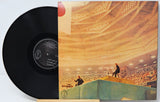 A vinyl record partially pulled from its illustrated sleeve, photographed on a plain light background. The record is black with a printed center label, and the sleeve shows a warm-toned interior scene with a large crowd and architectural ceiling details.