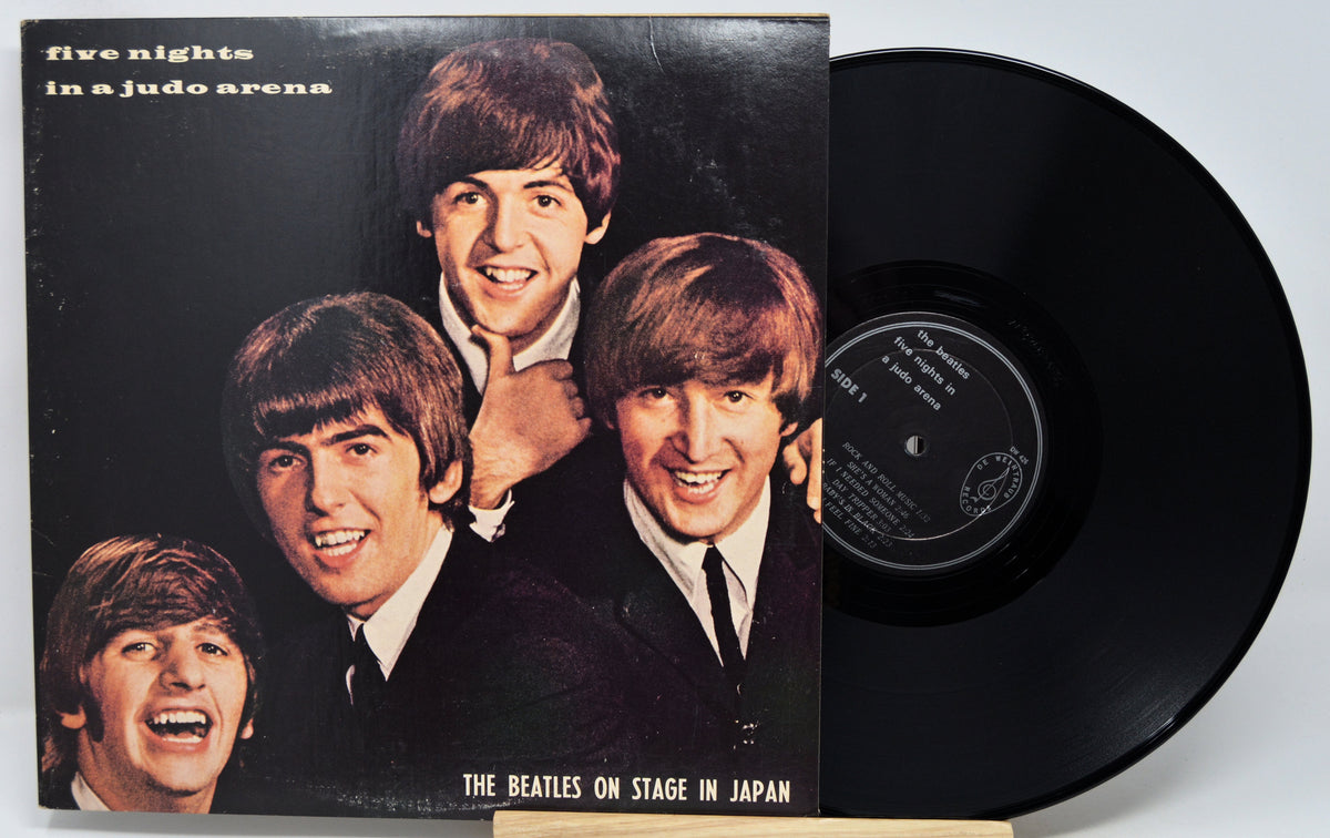 The Beatles 'Five Nights in a Judo Arena' vinyl record LP with its paper sleeve cover partially obscuring the black record, photographed against a light gray background.