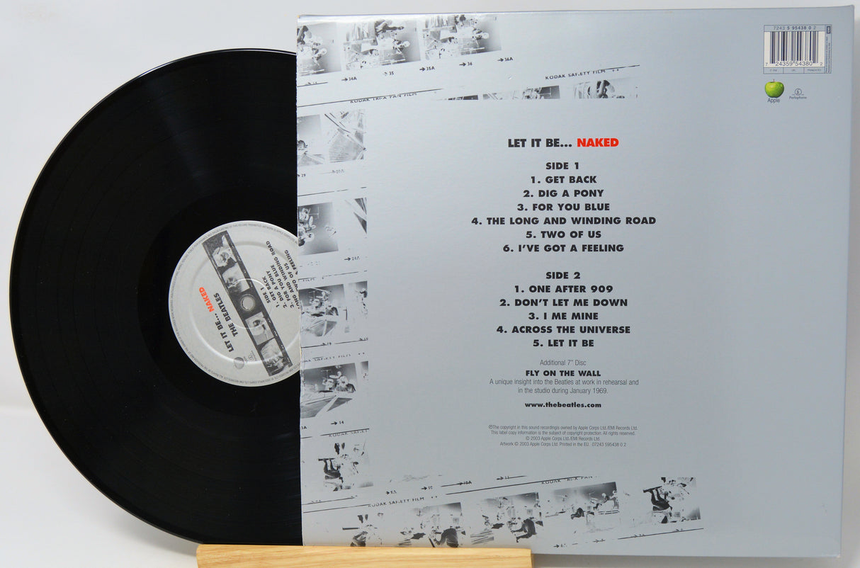 Vinyl record shown with its back cover standing behind it on a small wooden stand. The black LP is partially visible on the left, and the cover displays the track listing and printed artwork for the release 'Let It Be.