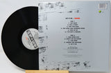 Vinyl record shown with its back cover standing behind it on a small wooden stand. The black LP is partially visible on the left, and the cover displays the track listing and printed artwork for the release 'Let It Be.