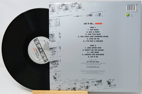 Vinyl record shown with its back cover standing behind it on a small wooden stand. The black LP is partially visible on the left, and the cover displays the track listing and printed artwork for the release 'Let It Be.