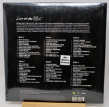 Back cover of a Beatles 'Live at the BBC' vinyl record sleeve, sealed in plastic shrink wrap, displayed upright on a small wooden stand against a light gray background.