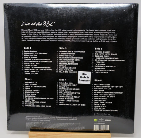 Back cover of a Beatles 'Live at the BBC' vinyl record sleeve, sealed in plastic shrink wrap, displayed upright on a small wooden stand against a light gray background.