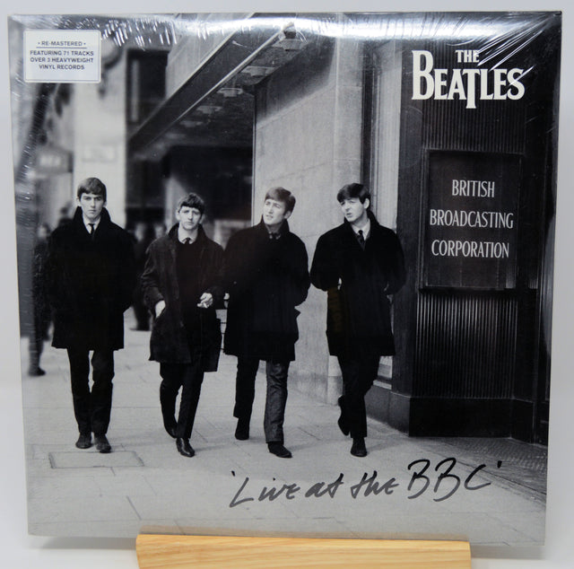 The Beatles 'Live at the BBC' vinyl record LP cover, sealed in its original plastic shrink wrap and displayed on a wooden stand. The black-and-white cover features the band members walking down a street, with 'The Beatles' and 'British Broadcasting Corporation' logos printed on the top right, and the handwritten title 'Live at the BBC' at the bottom.