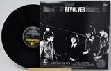 A black vinyl record partially pulled from its sleeve, showing the front cover for the band Revolver's 'Long Play 33 RPM'. The sleeve features a high-contrast black and white photo of the band members, with the band name 'REVOLVER' in bold white text at the top.