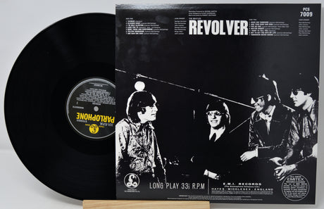 A black vinyl record partially pulled from its sleeve, showing the front cover for the band Revolver's 'Long Play 33 RPM'. The sleeve features a high-contrast black and white photo of the band members, with the band name 'REVOLVER' in bold white text at the top.