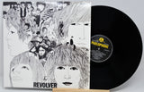The Beatles' 'Revolver' vinyl record LP and its illustrated cover sleeve displayed on a small wooden stand against a light gray background. The black vinyl record is partially visible behind the sleeve, with the Parlophone label visible.