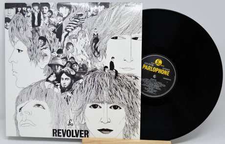 The Beatles' 'Revolver' vinyl record LP and its illustrated cover sleeve displayed on a small wooden stand against a light gray background. The black vinyl record is partially visible behind the sleeve, with the Parlophone label visible.
