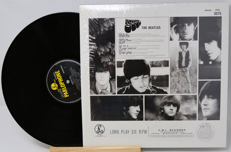 The Beatles 'Long Play 33 RPM' vinyl record LP and its outer paper sleeve, sealed in plastic shrink-wrap, displayed on a small wooden stand against a white background.