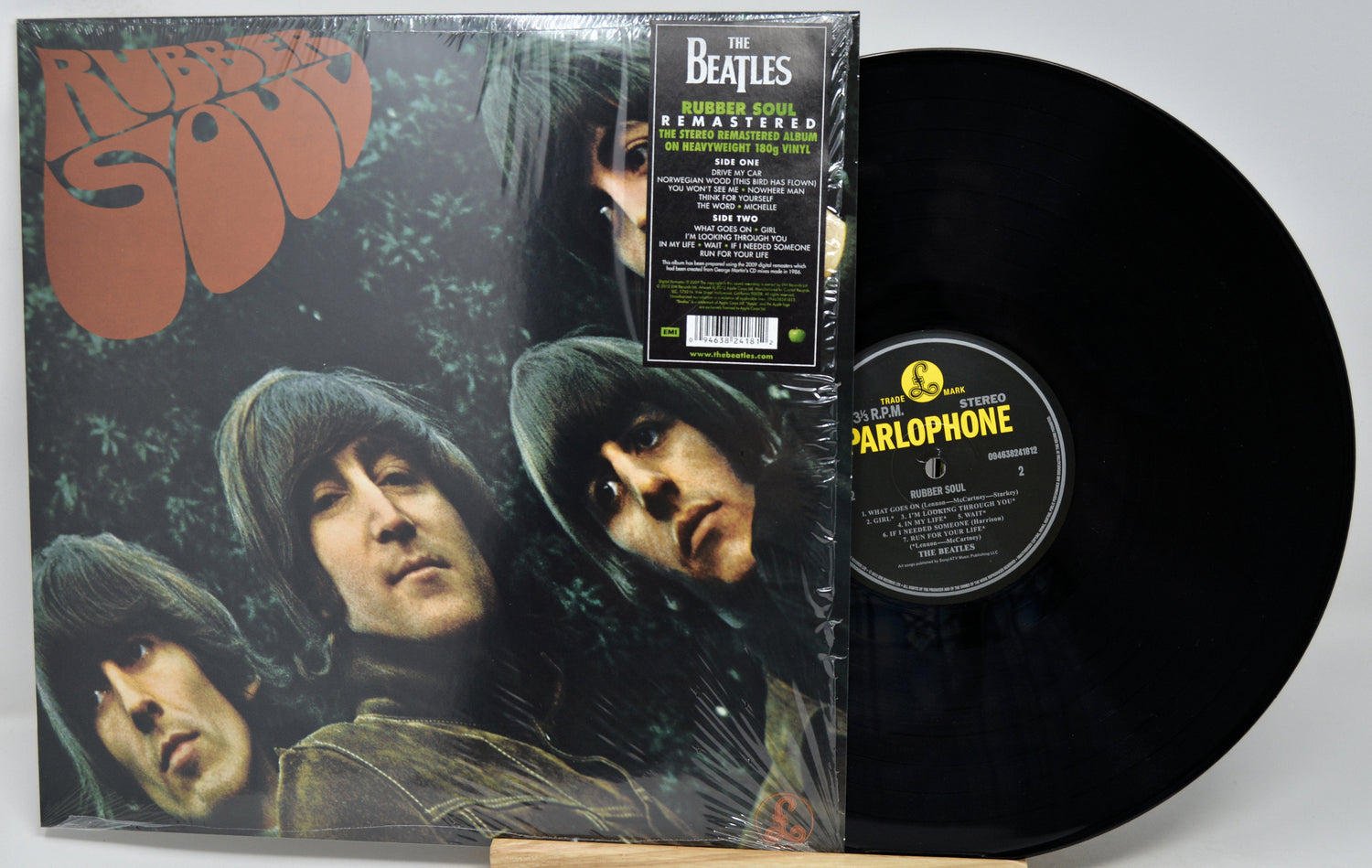 The Beatles - Rubber Soul remastered 12-inch vinyl record LP with its outer sleeve and a black vinyl record partially pulled out, displayed on a wooden stand against a light gray background.
