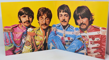 A vintage Beatles poster print showing the band members in colorful military-style jackets against a bright yellow background, photographed standing on a white surface.