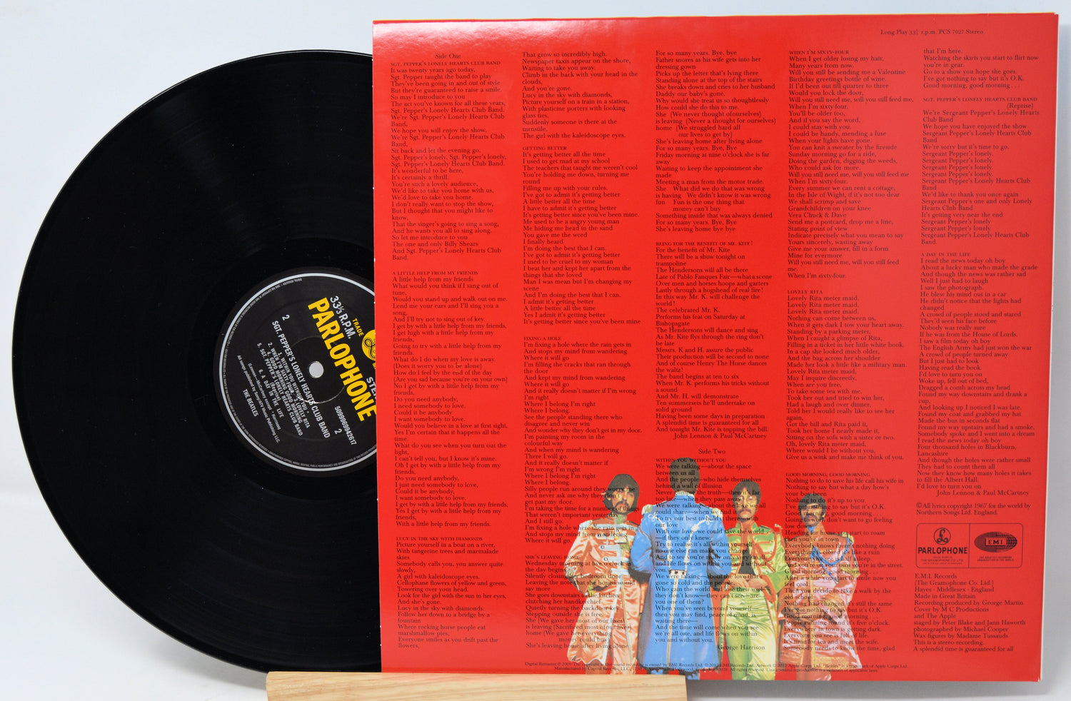 A black vinyl record partially pulled from its red back cover, which features dense printed text and a small illustrated group of four people near the bottom. The record label reads Parlophone.