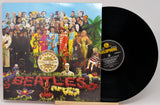 The Beatles 'Please Please Please' vinyl record LP and its illustrated cover jacket, shown on a light gray background. The black record is partially pulled from the sleeve, revealing the center label with 'Harlophone' branding.