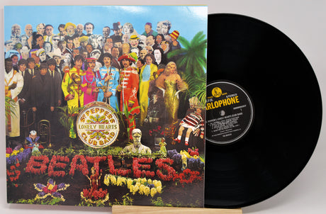 The Beatles 'Please Please Please' vinyl record LP and its illustrated cover jacket, shown on a light gray background. The black record is partially pulled from the sleeve, revealing the center label with 'Harlophone' branding.