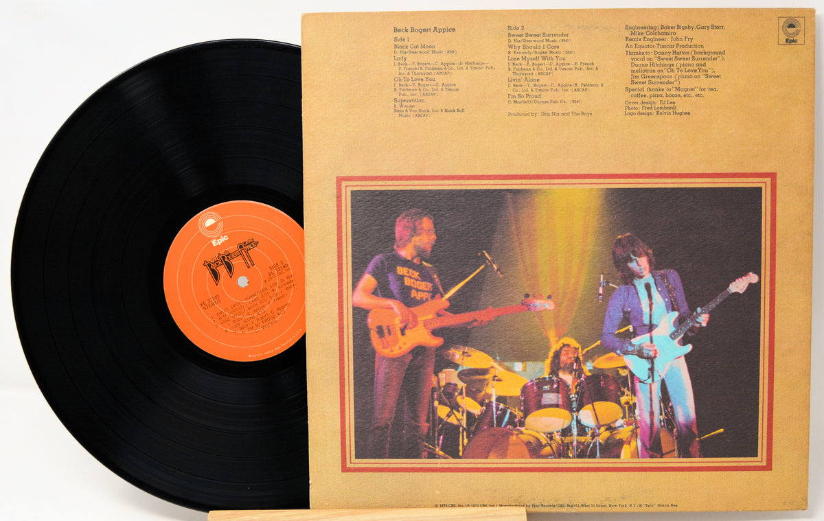 A vinyl record partially pulled from its sleeve, showing the black record with an orange label next to the tan printed album cover featuring a band photo and track listings.