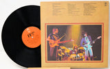 A vinyl record partially pulled from its sleeve, showing the black record with an orange label next to the tan printed album cover featuring a band photo and track listings.
