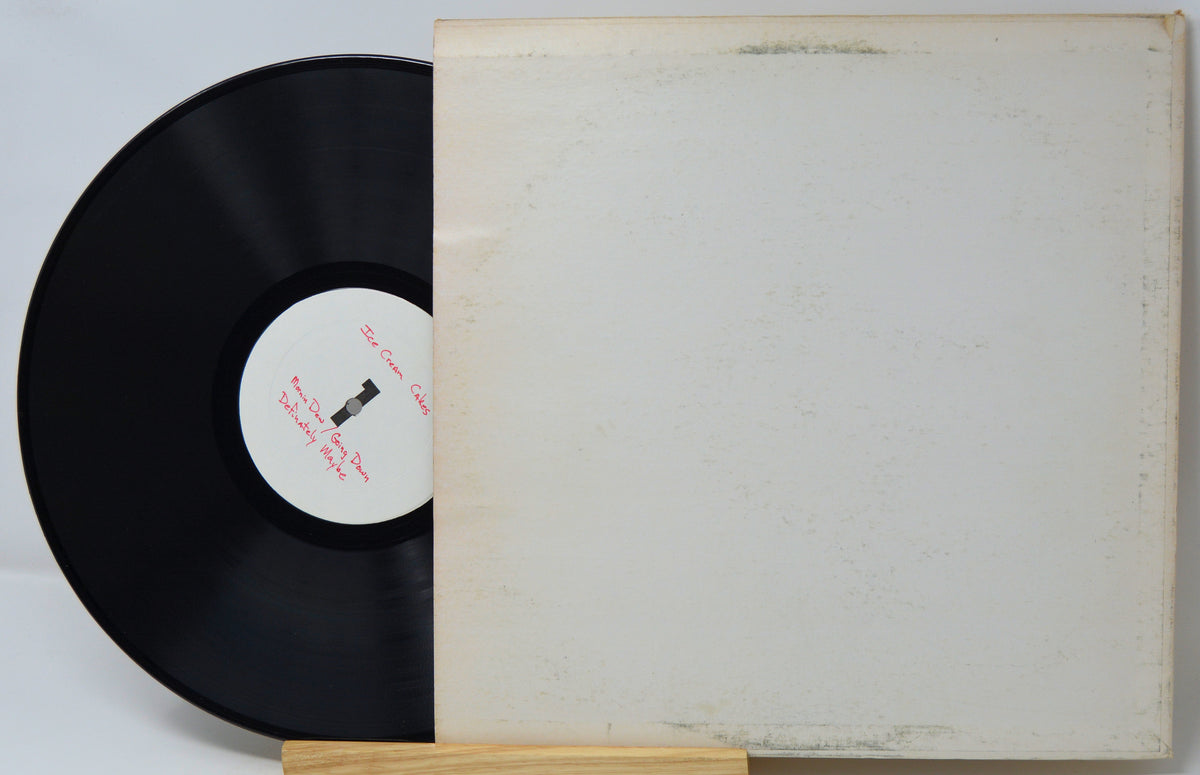 A vinyl record partially pulled from its sleeve, displayed on a small wooden stand against a plain light background. The sleeve is off-white with visible aging and scuffs, and the black record has a white center label with red text.