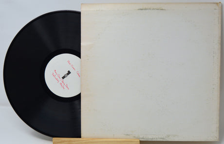 A vinyl record partially pulled from its sleeve, displayed on a small wooden stand against a plain light background. The sleeve is off-white with visible aging and scuffs, and the black record has a white center label with red text.