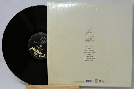 A vinyl record partially pulled from its cream-colored sleeve, which is still wrapped in clear plastic. The record is black with a gold-foil label, and the sleeve features a tracklist printed in black text.