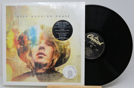 Beck's 'Morning Phase' vinyl record album in its shrink wrap, shown with the black LP partially pulled from the cover on a light gray background.