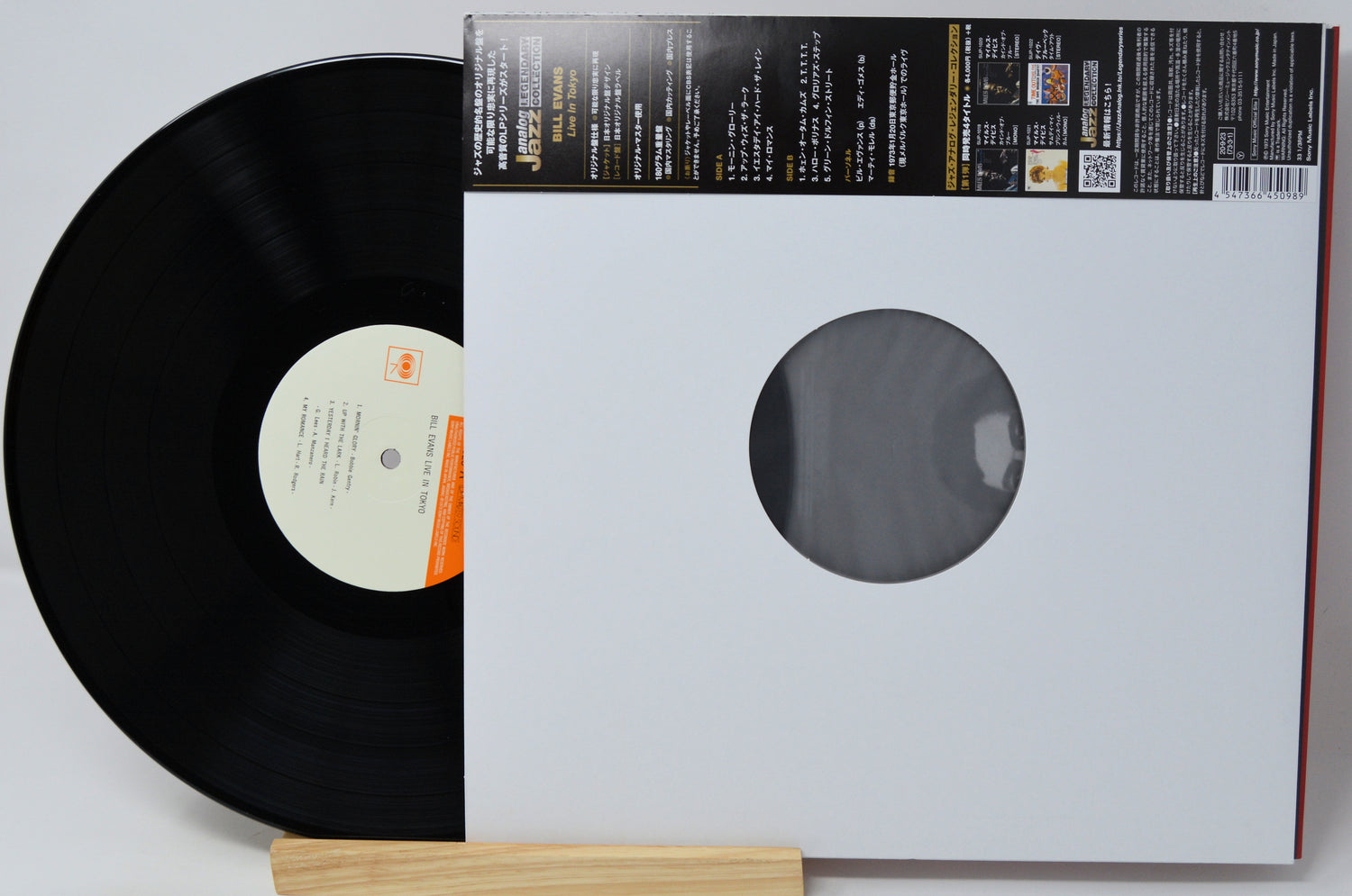 A vinyl record partially pulled from its white paper sleeve, with the black record visible on the left and the sleeve showing a circular window and printed track information on a light gray background.