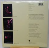 The back cover of a vinyl record sleeve, wrapped in protective plastic, displayed on a wooden stand. The cover is cream-colored with black text listing track titles and credits, alongside three vertical black-and-white photographs of a musician performing on stage.