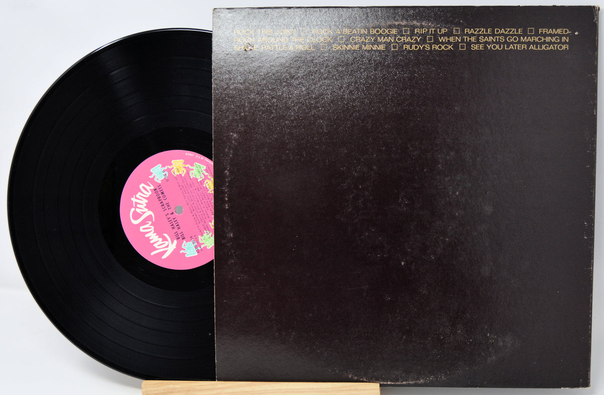 A vinyl record partially pulled from its sleeve, showing the black record and a pink center label next to the dark brown album cover with gold text.
