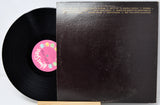 A vinyl record partially pulled from its sleeve, showing the black record and a pink center label next to the dark brown album cover with gold text.