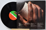 A vinyl record partially pulled from its album cover, showing the black record and the back cover artwork with a close-up of a hand holding a torn note. The record label has a green, white, and orange center label.