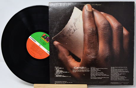 A vinyl record partially pulled from its album cover, showing the black record and the back cover artwork with a close-up of a hand holding a torn note. The record label has a green, white, and orange center label.