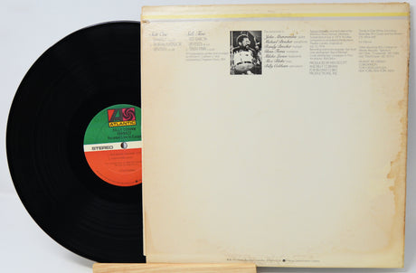 A vintage vinyl record partially pulled from its cream-colored paper sleeve, showing the black record, a green and orange center label, and printed tracklist text on the cover.