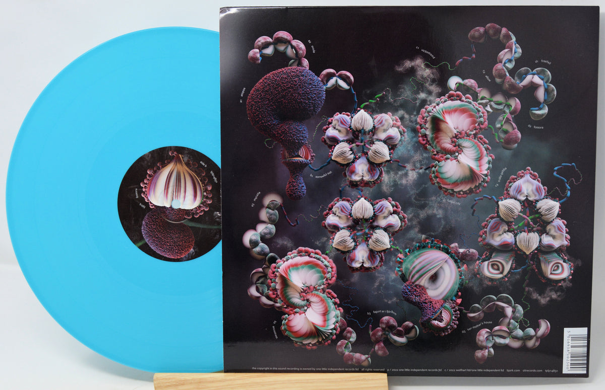 A vinyl record partially pulled from its album sleeve, displayed on a small wooden stand. The record is a bright translucent aqua blue, and the sleeve features a dark cover with surreal, organic floral and mushroom-like illustrations in pink, purple, and white.