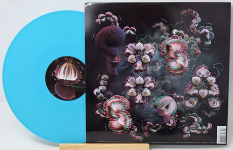 A vinyl record partially pulled from its album sleeve, displayed on a small wooden stand. The record is a bright translucent aqua blue, and the sleeve features a dark cover with surreal, organic floral and mushroom-like illustrations in pink, purple, and white.