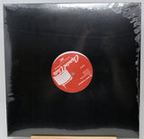 A black vinyl record sleeve or album cover wrapped in clear plastic, with a red circular label in the center featuring white text and graphics. The product is photographed against a light gray background with a small wooden stand visible at the bottom.