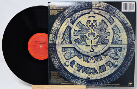 A vinyl record partially pulled from its sleeve, showing the black record with a bright orange-red center label next to the album cover featuring a large circular blue-and-gold emblem.