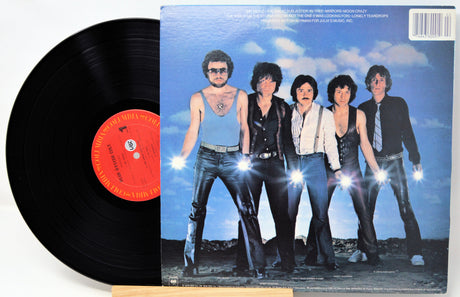 A vinyl record partially pulled from its album cover, showing the black record with a red-orange center label beside the printed cover art of a band standing against a blue sky.