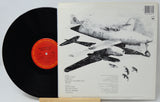 A vinyl record partially slid out of its printed album cover, showing a black record with a red-orange center label beside the back cover featuring black-and-white illustrations of military aircraft.