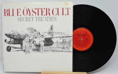 Blue Oyster Cult's 'Secret Treaties' vinyl record album, showing the illustrated white cover with red title text and a black LP partially pulled out, with a red center label.