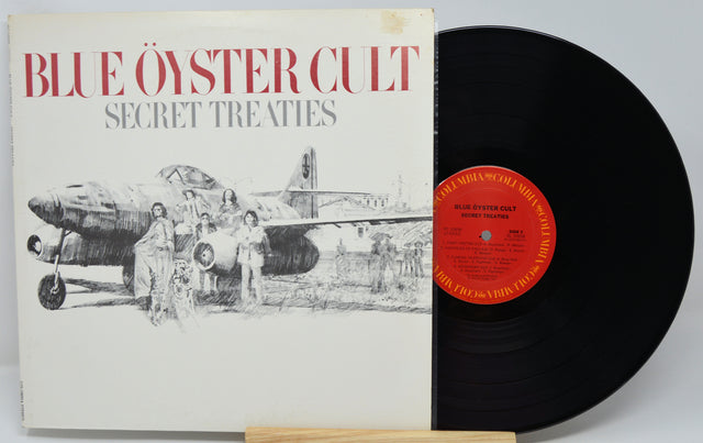 Blue Oyster Cult's 'Secret Treaties' vinyl record album, showing the illustrated white cover with red title text and a black LP partially pulled out, with a red center label.