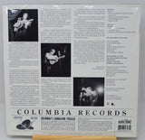 The back cover of a Columbia Lamination Records music release, featuring track listings, biographical text, and black-and-white photos of a performer on stage, sealed in a plastic sleeve.