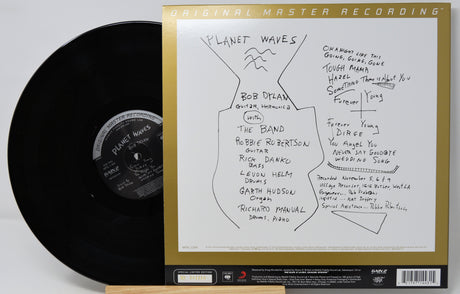 Bob Dylan's 'Planet Waves' vinyl record album shown with the black LP partially pulled from the gatefold cover, which is signed and features tracklist handwriting on a white background.