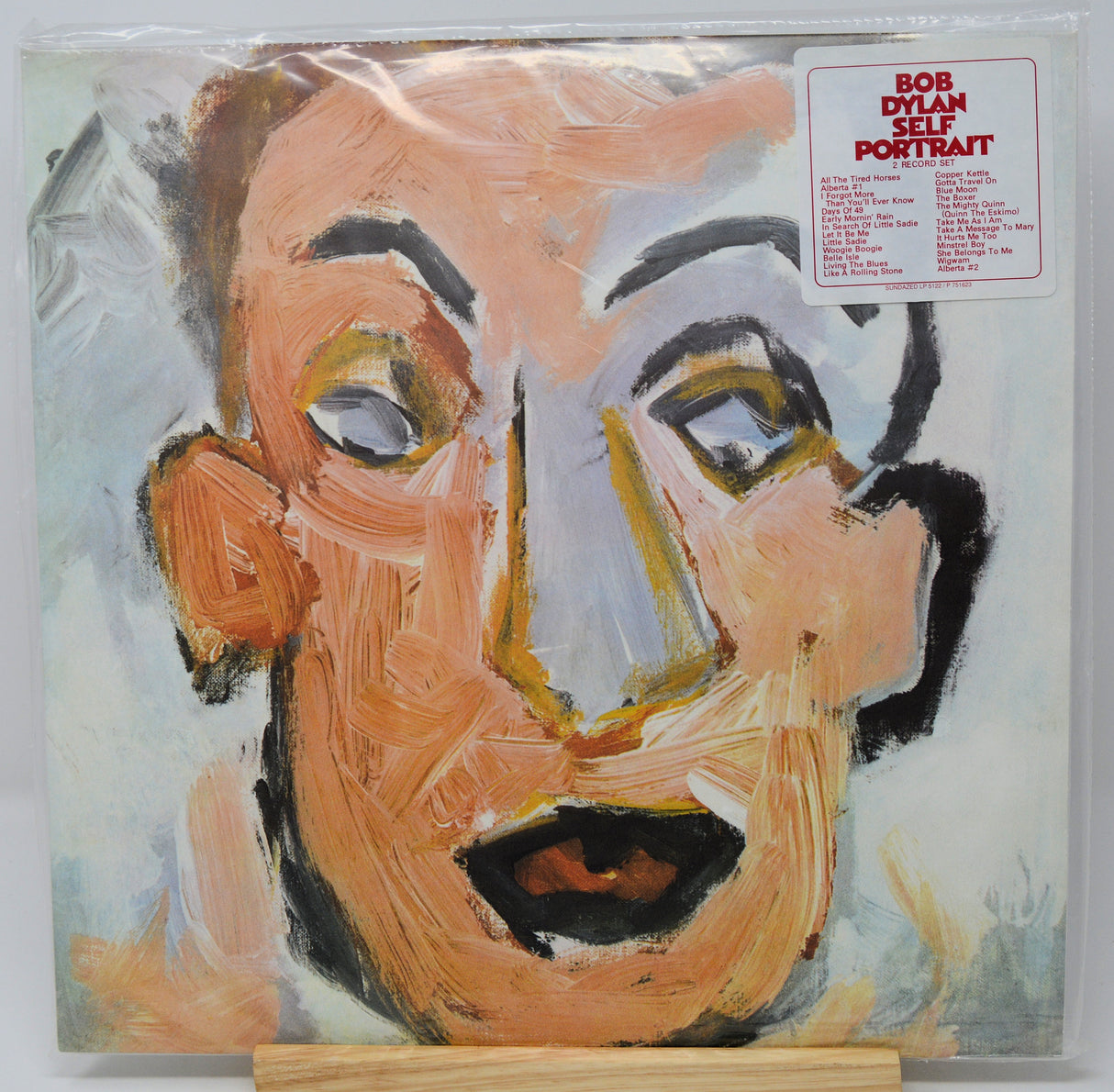 Bob Dylan 'Self Portrait' vinyl record LP cover inside a clear plastic shrink wrap, displayed on a small wooden stand against a white background.