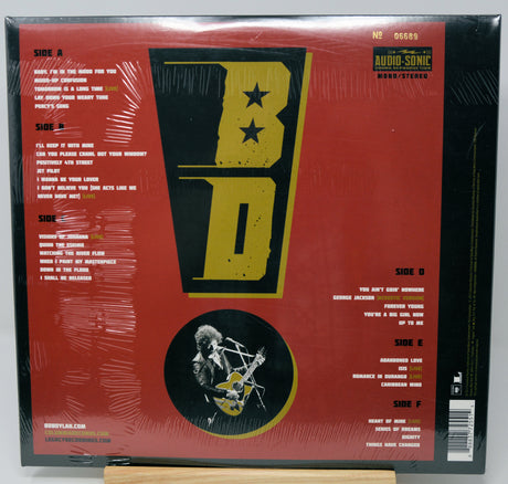 The back cover of a vinyl record album wrapped in protective plastic, featuring a bold red background with a large black and yellow 'BD' logo, track listings, and an illustration of a musician playing guitar.