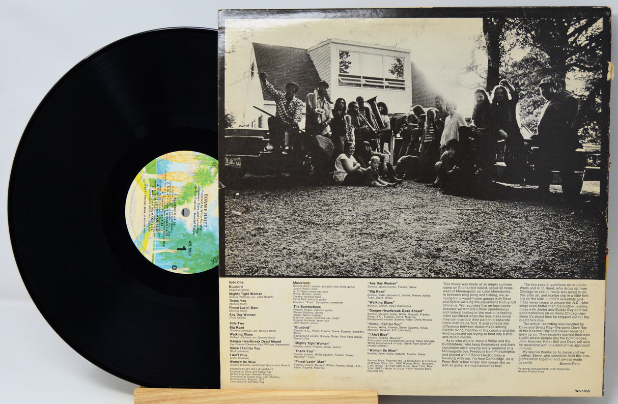 A vinyl record partially pulled from its sleeve, showing the black record on the left and the back cover of the album on the right with a black-and-white group photograph and printed track text on a cream background.