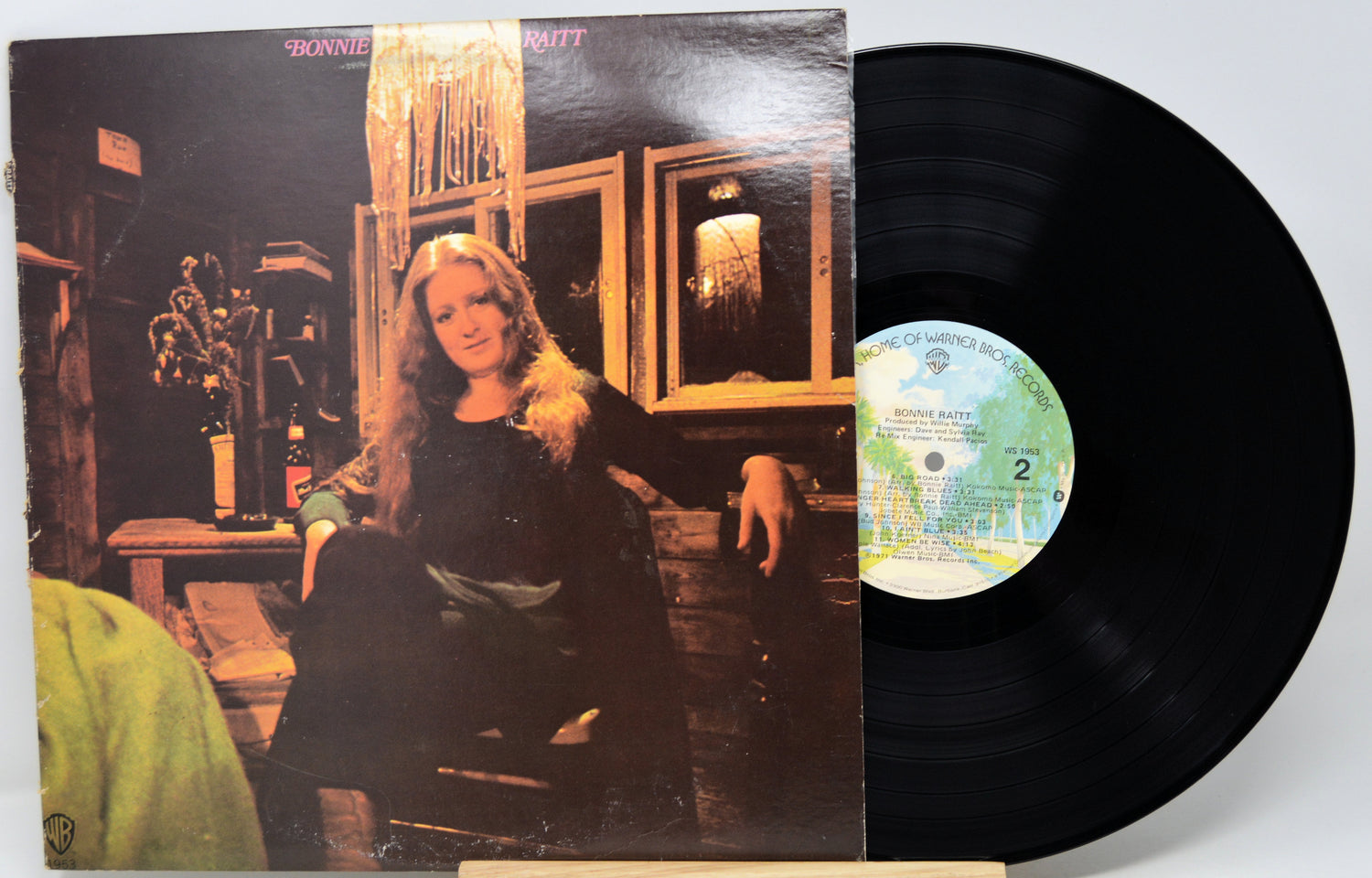 A vinyl record partially pulled from its album sleeve, showing the black record and the printed cover art of a woman in a dim interior. The center label is visible, and the background is plain white.
