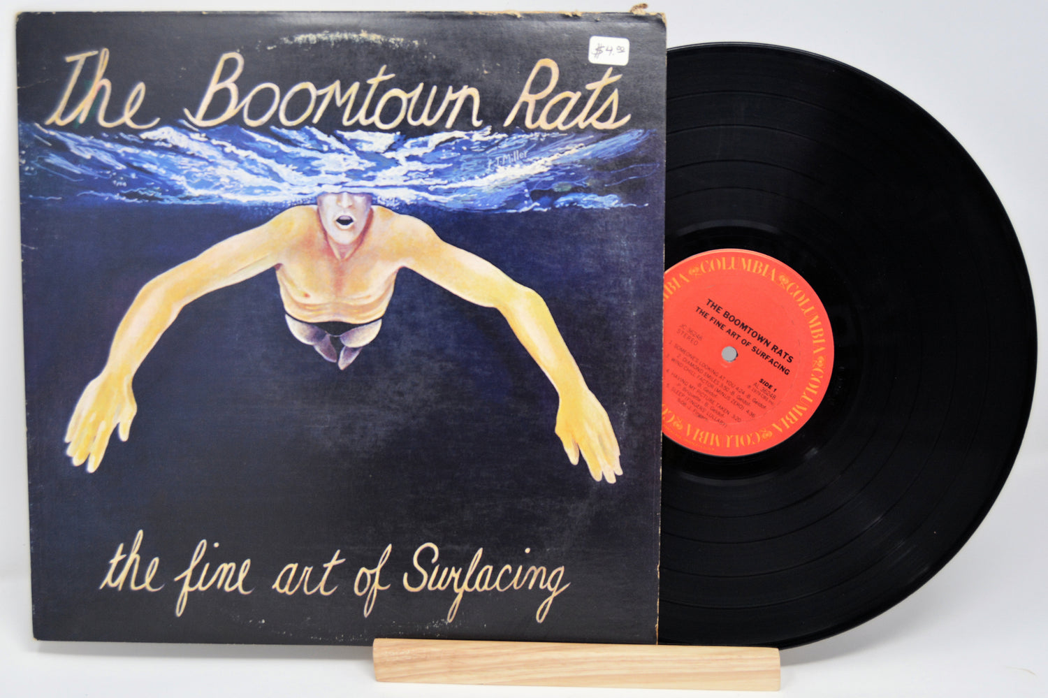 A vinyl record titled 'The Boomtown Rats - The Fine Art Of Surfacing' with the image of a person swimming on the cover, placed next to its sleeve.