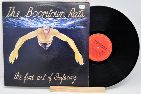 A vinyl record titled 'The Boomtown Rats - The Fine Art Of Surfacing' with the image of a person swimming on the cover, placed next to its sleeve.