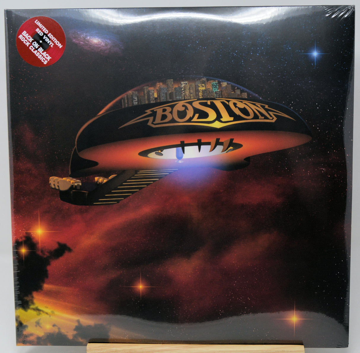Boston band album cover for 'Boston' on a vinyl record, showing a futuristic flying saucer over a dramatic red and orange sky with stars. A red circular sticker in the top-left corner reads '12" VINYL LP NEW BRITAIN CD RELEASE'.